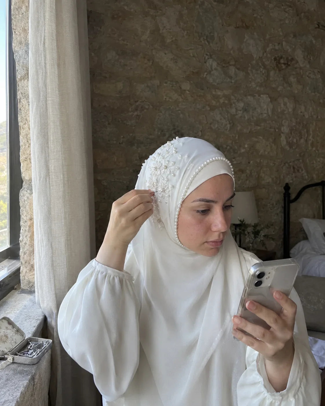 Femme qui fixe l'épingle de son hijab de mariage blanc perlé au-dessus de l'oreille, en se regardant dans son téléphone, avant la fête