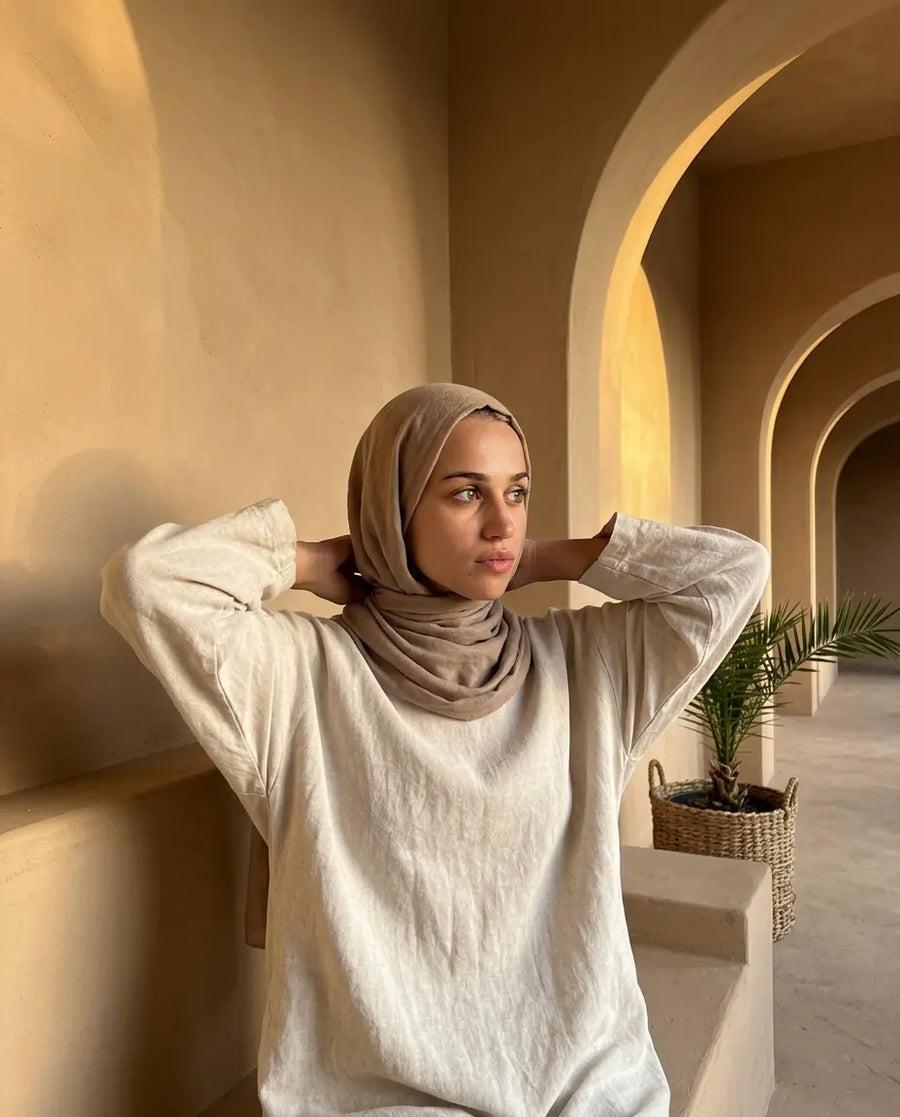 Femme qui noue son hijab derrière la nuque, sous une enfilade d'arcades en terre, au soleil couchant