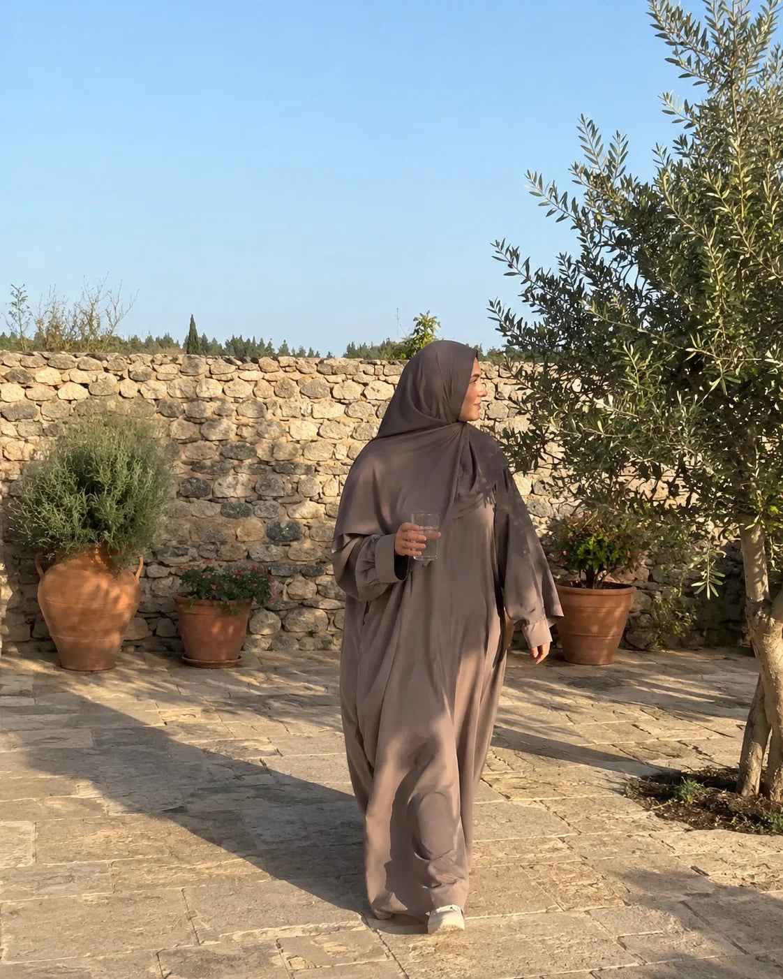 Femme en hijab et abaya qui traverse une cour ensoleillée en été, un verre d'eau à la main