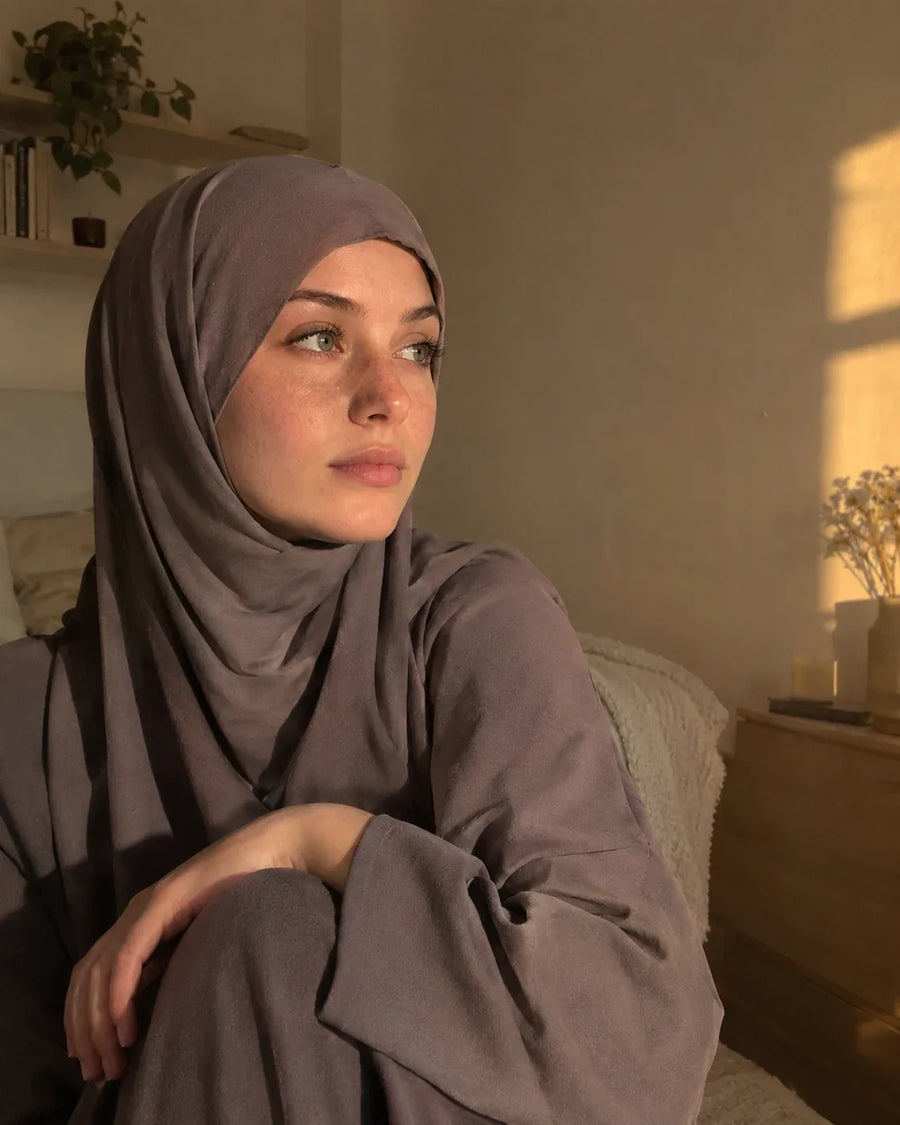 Femme en hijab mauve mat, assise dans une chambre au soleil du soir, le regard tourné vers la fenêtre