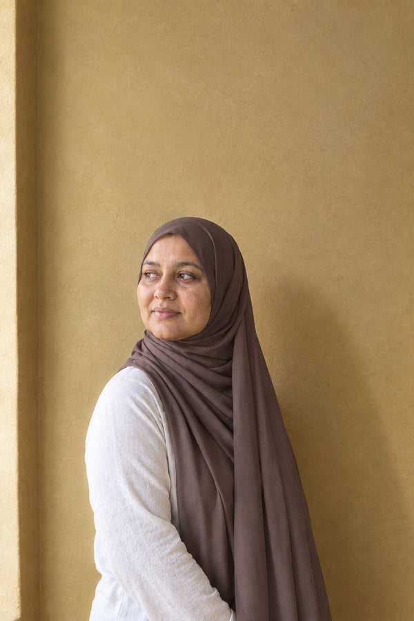 Femme portant le foulard hijab mousseline taupe, noué, devant un mur ocre